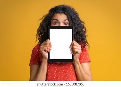 Cute funny girl hiding half of her face behing the tablet with blank white screen making open-eyed face expression. Isolated on bright colored yellow background. Mock up, copy space for your text. - Powered by Shutterstock