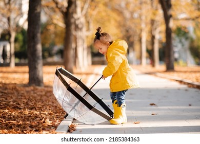 Cute Funny Kid Girl 2-3 Year Old Wear Yellow Rain Jacket And Boots Hold Umbrella In Autumn Park Outdoor Over Fallen Leaves. Fall Season. Childhood. 