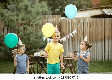 Cute Funny Kids Children Celebrating Birthday With Family Or Friends In A Backyard. Birthday Party. Kid Wearing Party Hat And Launch Balloons Into The Sky