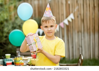 Cute Funny Nine Year Old Boy Celebrating His Birthday With Family Or Friends In A Backyard. Birthday Party. Kid Wearing Party Hat And Holding Gift Box