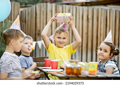 Cute Funny Nine Year Old Boy Celebrating His Birthday With Family Or Friends In A Backyard. Birthday Party. Kid Wearing Party Hat And Holding Gift Box