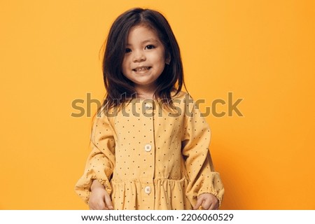 a cute, funny preschool girl stands on an orange background with an empty space for an advertising mockup in a summer dress and makes a funny emotional face
