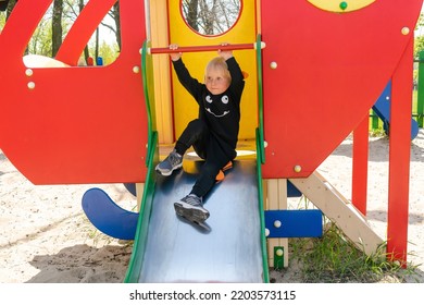 Cute Funny Smiling Blonde Little Young Toddler Kid Child Boy Going Down Slide In Playground. Children Physical,emotional Development And Childhood Daycare, Kindergarten Concept.