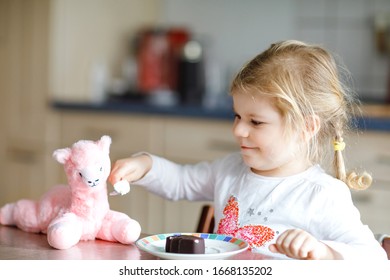 Cute Funny Toddler Girl Eating Chocolate Ice Cream At Home. Happy Healthy Baby Child Feeding Plush Lama Toy With Sweet Ice-cream. Lovely Kid Enjoying Dessert Pudding.