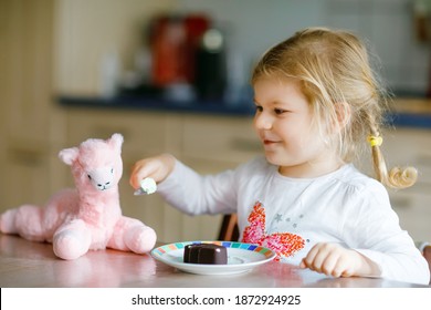 Cute Funny Toddler Girl Eating Chocolate Ice Cream At Home. Happy Healthy Baby Child Feeding Plush Lama Toy With Sweet Ice-cream. Lovely Kid Enjoying Dessert Pudding.