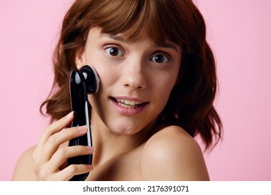 a cute, funny woman with a funny facial expression stands on a pink background and massages her facial skin with an electric black massager. Horizontal photography in the studio