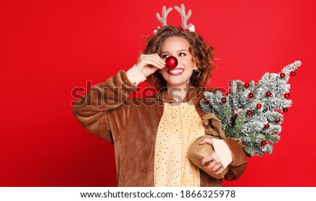 cute funny young woman in a Christmas reindeer costume  with red bauble and Christmas tree on colorful red background
