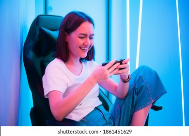 Cute gamer girl sit on a gaming chair, smiling and playing mobile online game on a smartphone. Having fun playing with friends online.