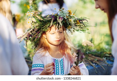 Cute Girl Blonde In White National Dress Weaves A Wreath Of Flowers And Grass At The Pagan Festival Of Spring