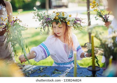 Cute Girl Blonde In White National Dress Weaves A Wreath Of Flowers And Grass At The Pagan Festival Of Spring