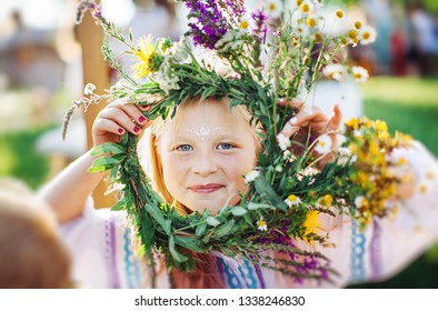 Cute Girl Blonde In White National Dress Weaves A Wreath Of Flowers And Grass At The Pagan Festival Of Spring