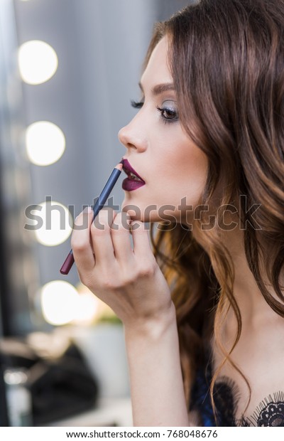 Cute cute girl in a blue dress painted at the\
mirror, putting on lip pencil professional makeup, makeup, profile,\
close-up
