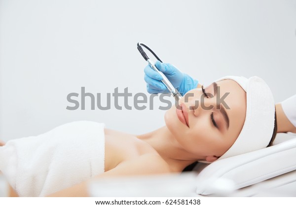 Cute girl\
with brown hair fixed behind,clean fresh skin naked shoulders\
wearing white bath robe and hair bandage, doing cosmetic procedure\
at light medical background,\
microdermabrasion.