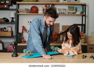 Cute girl checking wheels of blue skateboard after fixing them with help and advice of her father while both standing by wooden table - Powered by Shutterstock