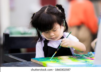 Cute Girl Is Coloring On A Canvas. Children Work On Watercolor Art. Child Wear Black Aprons To Prevent White School Uniforms From Messing Up The Color. Kid Sit On A High Chair.