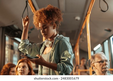 Cute girl with curly hair standing on the bus, holding onto a handrail and looking down at her phone in her left hand. - Powered by Shutterstock