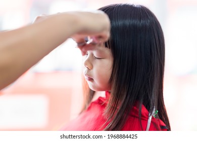 Cute Girl Cut The Bangs. The Barber's Hand Held Scissors Skillfully Cutting The Cutie Kid. Child Is Covered With A Red Veil. A 4 Year Old Children Sitting In A Beauty Salon.