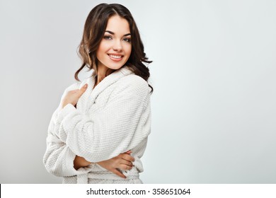 Cute girl with dark hair, big eyes and dark eyebrows  wearing white bath robe, looking at camera and smiling, hugging, model with light nude make-up, white studio background, beauty photo, copy space