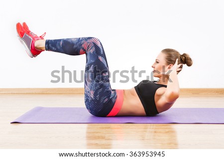 Cute girl with dark hair wearing pink snickers, dark leggings and black short top doing reverse crunch at gym, fitness, white wall and wooden floor.