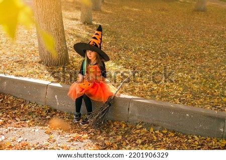 A cute girl dressed up in a witch costume is sitting on the outskirts of the sidewalk, covered with dry autumn foliage with a carved pumpkin, waiting for a traditional hike on Halloween evening