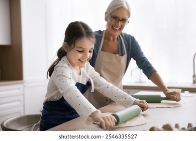 Cute girl flattening dough with rolling-pin helps with cooking process to 60s granny, wear aprons preparing holiday family recipe pie in modern kitchen. Multi-generational relatives joint activity - Powered by Shutterstock