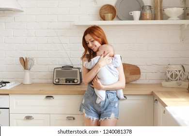 Cute girl with ginger hair embracing adorable baby in blue bodysuit, cradling him sleep to music on radio, standing at kitchen counter and smiling. Pretty sister babysitting her infant niece