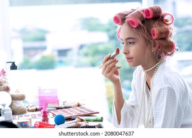 Cute girl with hair curlers doing makeup on her face, looking in the mirror at  the makeup table,  brushes for makeup in the hands