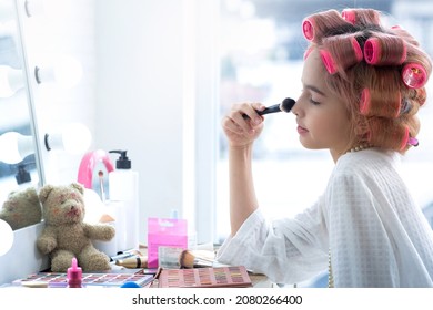Cute girl with hair curlers doing makeup on her face at  the makeup table, using makeup brush