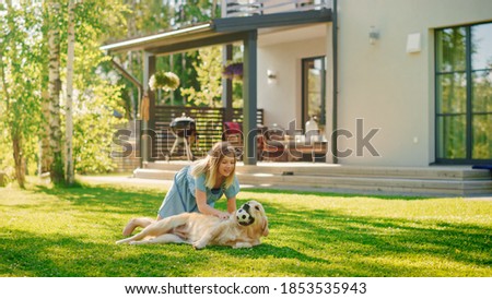 Cute Girl Has fun with Happy Golden Retriever Dog on the Backyard Lawn. She Pets, Play, Tackle it on the Ground And Scratches Back. Happy Dog Plays with Toy Ball. Idyllic Summer House.