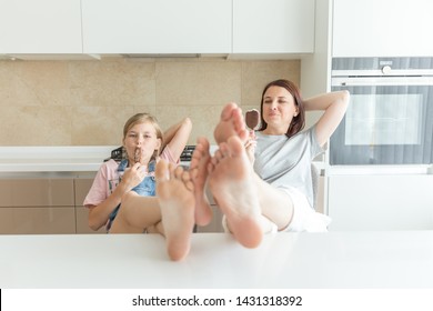 Cute Girl And Her Mother Are Smiling While Eating Ice Cream In The Kitchen With Legs On A Table