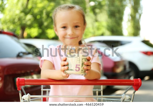 Cute girl holding bank with money in hands and\
sitting in shopping cart,\
outdoors