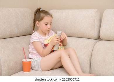 Cute Girl At Home On The Couch With A Big Glass Of Popcorn And Coca Cola Trying To Figure Out The Remote Control To Switch Tv Channels.