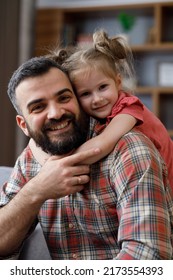 Cute Girl Hugging Her Beloved Dad Around Neck. Young Handsome Father With Cute Daughter Sitting On A Couch, Looking At Camera And Smiling.Family Love And Family Bond Concept. Happy Carefree Childhood