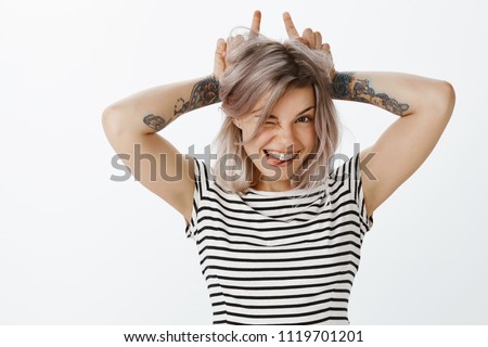 Cute girl little devilish inside. Portrait of cute playful woman with fair hair, braces and tattoos, showing horns with index fingers on head, winking and smiling while sticking out tongue