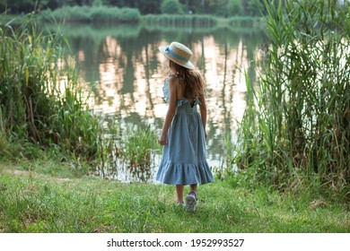 A Cute Girl In A Long Blue Dress And Straw Hat Stands On The Shore Of The Lake. Back Of A Child In The Public Park With Water, Bushes, Green And Dried Grass In Summer. Young Beautiful Lady On Walking.