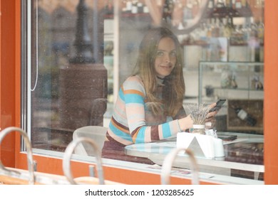 Cute Girl With Long Hair Sitting In Caffe And Chatting On Mobile Phone. Photo Taken From Outside, With A See Through The Window. 