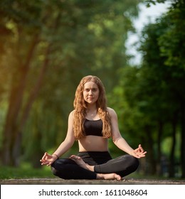 Cute Girl Meditating In The Lotus Position, Padmasana Asana On The Background Of A Blurred Green Park Under The Sun's Rays On A Warm Day. Morning Yoga Training Outdoor All Day Energy Boost