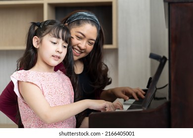 Cute Girl And Mother Playing The Piano Together In Living Room At Home. Asian Pianist Teacher Teaching Little Girl Kid Student To Play Piano At School.