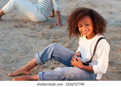 Cute girl with old-fashioned camera. Female model with curly hair and in white pullover holding camera on beach. Mother in background. Childhood, hobby concept - Powered by Shutterstock