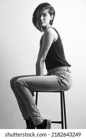 Cute Girl In Pixie Hair, Wearing Top And Jeans And Short Hair Sitting On Chair. 