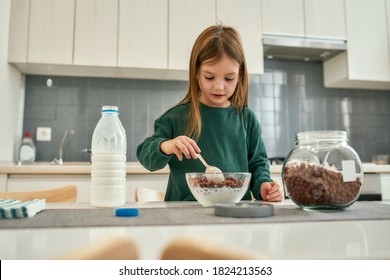 A cute girl prepearing quick breakfast made of chocolate cereal balls and milk stirring with a spoon in a huge light kitchen at home
