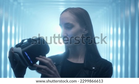 Cute Girl puts a virtual reality headset or helmet on her eyes, enjoys the experience. Looks inside the glass of augmented reality glasses. Neon light in the studio. Digital art, immersion.