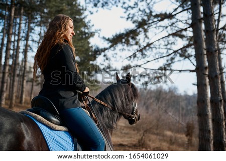 A cute girl riding a horse in the forest background looks in front of herself.