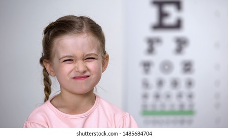 Cute Girl Screwing Up Her Eyes, Trying To See Letters On Table For Vision Test