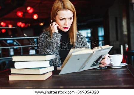 Cute girl with short light hair and red lips wearing black and gray sweater sitting in cafe with cup of coffee and opened book, holding pan.