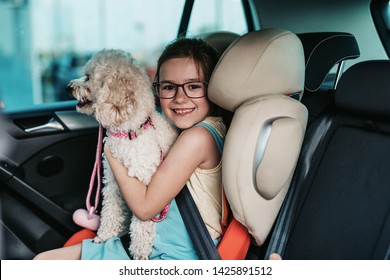 Cute Girl Sitting In A Car With Her Dog On A Safety Child Car Seat.