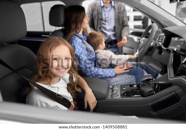 Cute\
girl sitting in new big automobile, looking at camera and smiling\
in auto showroom. Pretty child with parents and brother testing and\
examining cars in auto salon. Concept of\
buying.