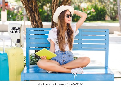 Cute girl sitting on a blue bench and playing on a tablet in yellow case. She wears denim shorts, white t-shirt, dark sunglasses, white sneakers and straw hat. She sits with two suitcases next to her. - Powered by Shutterstock