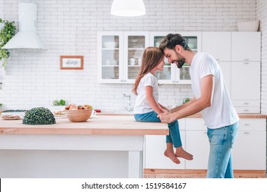 cute girl sitting on the table and father standing on the kitchen and looking at each other, side view