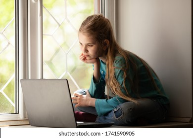 Cute girl sitting on the window sill with laptop studying online at home. Remote education concept. 